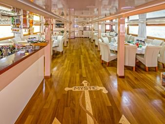 Innenraum des MY Mystic Salons mit Holzboden und Anker-Intarsie, weißen Essstühlen, Buffettheke und großen Fenstern.