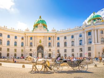 Fiaker im Innenhof der Alten Hofburg in Wien mit ihrer charakteristischen grünen Kuppel und cremefarbenen Barockfassade.