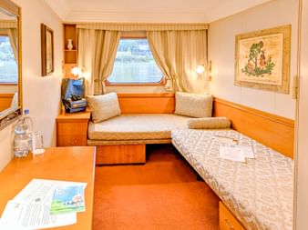 Kompakte Kabine auf der MS Prinzessin Katharina mit zwei Betten, Fenster mit Vorhängen, Holzmöbeln und orangem Teppich. Flussblick durch Fenster.