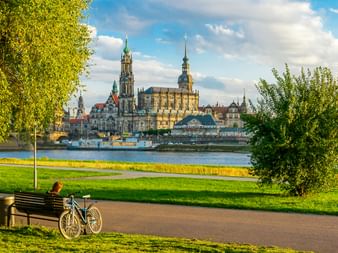 Fahrrad an Bank am Dresdner Elbufer. Historische Dresdner Skyline mit Kathedrale und Kirchtürmen über dem Fluss unter blauem Himmel.