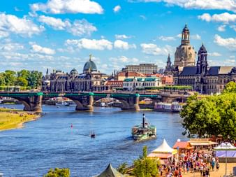 Dresdner Stadtbild mit Elbe, historischer Brücke und barocker Skyline mit Frauenkirche-Kuppel. Flussufer mit Besuchern und Booten sichtbar.
