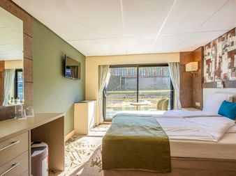 Moderne Schiffskabine auf der MS Lisabelle mit Doppelbett, olivgrünen Akzenten, Schreibtisch, TV und großem Fenster mit Vorhängen zum Deck.