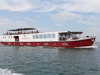MS Ave Maria Flusskreuzfahrtschiff mit rotem Rumpf und weißem Aufbau auf ruhigem Wasser unter bewölktem Himmel. Passagiere auf Oberdeck sichtbar.