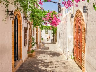 Sonnige Gasse in der Altstadt von Lindos mit weißen Wänden, Holztüren mit dekorativen Rahmen und rosa Bougainvillea-Blüten.