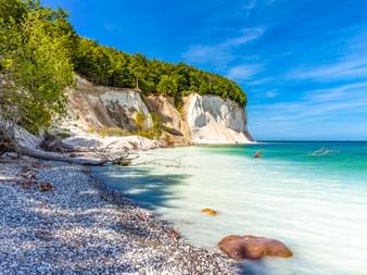 Weiße Kreidefelsen von Rügen mit grünem Buchenwald, Kieselstrand und türkisfarbenem Ostseewasser unter blauem Himmel.