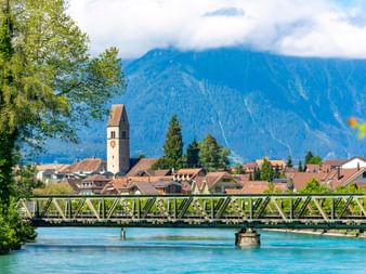 Blick auf Interlaken mit Kirchturm, Metallbrücke über die türkisfarbene Aare und Bergkulisse, die teilweise von Wolken bedeckt ist.