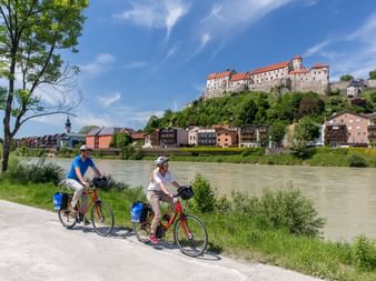 Zwei Radfahrer mit roten Rädern und blauen Taschen fahren auf einem Flussweg mit der Burg Burghausen auf einem Hügel und bunten Stadthäusern darunter.