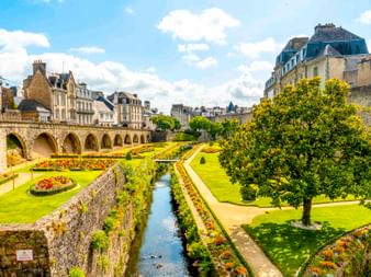 Formal gardens with colorful flowerbeds and water canal alongside ancient stone walls and arched bridge in Vannes, with historic buildings beyond.