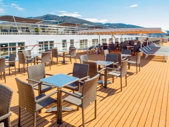 Sonnendeck der MS Prinzessin Katharina mit Holzboden, Rattanmöbeln, gestreiften Liegestühlen und Bergblick unter blauem Himmel.