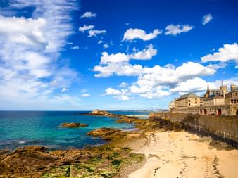 Sandstrand mit Felsformationen neben historischen Steinbefestigungen in Saint-Malo. Türkisfarbenes Meer und blauer Himmel mit weißen Wolken.