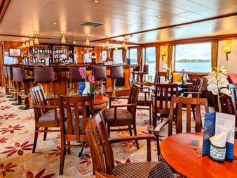 Innenraum der MS Rigoletto Lounge mit Holzbar, Esstischen, Stühlen und großen Fenstern mit Blick auf einen Fluss mit grünem Ufer.
