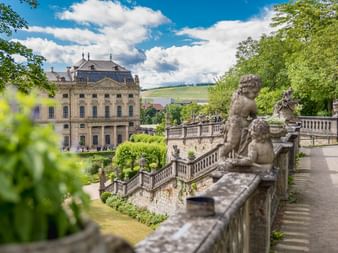 Steinbalustrade mit barocken Löwenstatuen im Würzburger Schlossgarten. Historisches Schlossgebäude im Hintergrund unter blauem Himmel mit Wolken.