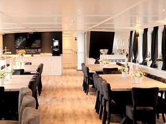 Elegantes Restaurant an Bord der MS Magnifique III mit weißen Tischen, dunklen Stühlen, Bogenfenstern mit schwarzen Vorhängen und Weinregal.