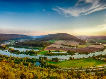 Luftaufnahme der Altmühl, die bei Riedenburg eine Hufeisenschleife bildet, mit grünen Feldern, bewaldeten Hügeln und blauem Himmel bei Sonnenuntergang.