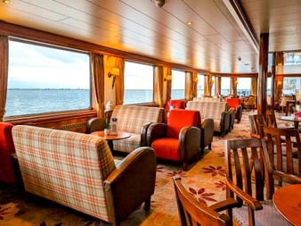 Innenlounge der MS Rigoletto mit karierten Sofas, roten Sesseln und Holzesstischen. Große Panoramafenster bieten Wasserblick.