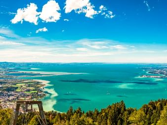 Luftaufnahme vom Pfänder mit Blick auf den Bodensee mit türkisfarbenem Wasser, umliegenden Städten und bewaldeten Hängen unter blauem Himmel.