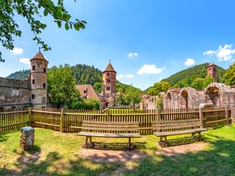 Historische Ruine Kloster Hirsau mit Steintürmen und Bogenmauern, Holzbänke im Vordergrund, umgeben von grünen Hügeln im Schwarzwald.