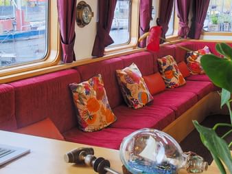 Heller Loungebereich auf der MS Fleur mit rot gepolsterten Sitzbänken, bunten Kissen, großen Fenstern mit bordeauxfarbenen Vorhängen und Grünpflanzen.