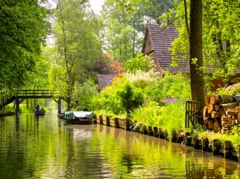 Holzhaus mit Ziegeldach neben einem ruhigen Kanal im Spreewald. Üppige grüne Vegetation säumt das Wasser, Boote und eine Holzbrücke sind sichtbar.