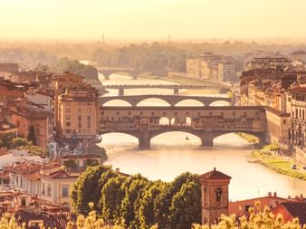 Aerial view of Florence showing the Ponte Vecchio and multiple bridges crossing the Arno River at sunset, with historic buildings and golden light.