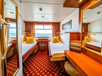 Standard-Doppelkabine auf der MS Rigoletto mit zwei Einzelbetten, gelben Kissen, Fenster mit Wasserblick, gemustertem Teppich und Holzmöbeln.