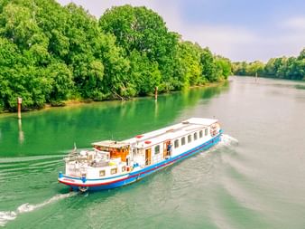 MS L'Estello, ein weißes Flusskreuzfahrtschiff mit blauen und roten Streifen, fährt auf einem grünen Fluss gesäumt von dichten Bäumen.