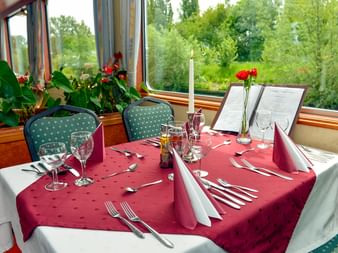 Restaurant an Bord der MS Fluvius mit Tischen, die mit burgunderfarbenen Tischdecken, Weingläsern und Kerzen gedeckt sind. Große Fenster zeigen grüne Ufervegetation.