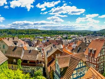 Panoramablick auf die Marburger Altstadt mit traditionellen Fachwerkhäusern, Ziegeldächern und grünen Hügeln unter blauem Himmel mit weißen Wolken.
