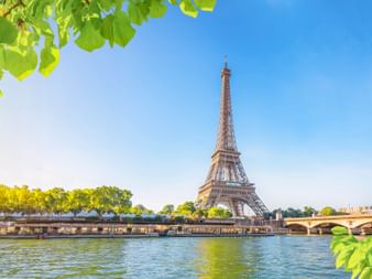 Eiffelturm über der Seine in Paris an einem sonnigen Tag, eingerahmt von grünem Laub mit Brücke und baumgesäumtem Flussufer.