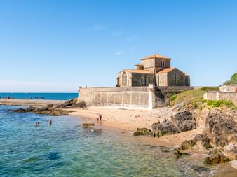 Prieuré Saint-Nicolas church on rocky coastline in Sables-d'Olonne. Stone building with terracotta roof overlooks sandy beach and blue Atlantic Ocean.