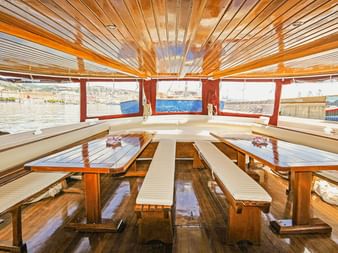 Innenraum des Schiffs MS Sveti Vid mit Holzbänken, Tischen und Lamellendecke. Große Fenster bieten Panoramablick auf den Hafen mit Booten.