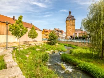 Bach fließt durch Bad Staffelstein mit historischem Steinturm und bunten Häusern. Grüne Vegetation säumt die Ufer unter blauem Himmel.