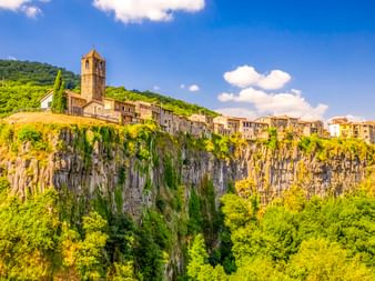Dorf Castellfollit de la Roca in Katalonien auf dramatischen Basaltklippen mit Kirchturm, umgeben von grünen Wäldern unter blauem Himmel.