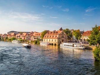 Historische Fachwerkhäuser entlang der Regnitz in Bambergs Klein Venedig mit Ausflugsbooten am Ufer unter blauem Himmel.