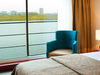Helle Kabine auf MS SE-Manon mit Doppelbett, großem Panoramafenster mit Flussblick, türkisfarbenem Sessel, Schreibtisch und warmer Beleuchtung.