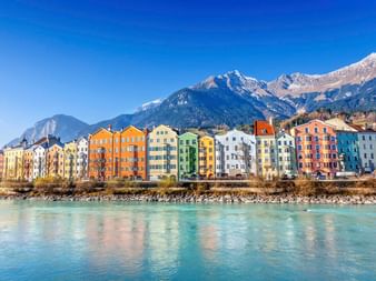 Reihe bunter historischer Gebäude am Inn in Innsbruck mit schneebedeckten Alpengipfeln im Hintergrund unter klarem blauen Himmel.