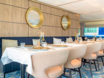 Elegantes Restaurant auf der MS Lisabelle mit langer Tafel, weißen Tischdecken, beigen Stühlen, holzgetäfelten Wänden mit goldenen Spiegeln und blauen Sitzgelegenheiten.