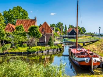 Traditionelles braunes Segelboot am ruhigen Kanal in Enkhuizen, mit historischen niederländischen Häusern mit roten Ziegeldächern und grünen Bäumen am Ufer.