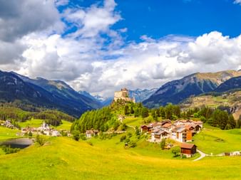 Schloss Tarasp auf einem bewaldeten Hügel über dem Dorf Scuol mit grünen Wiesen. Alpenberge und dramatische Wolken rahmen die Tallandschaft.