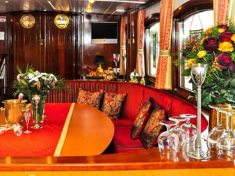 Eleganter Salon an Bord der MS Miro mit rot gestreifter Polsterung, poliertem Holz, Bullaugen und gedecktem Tisch mit Blumen und Gläsern.