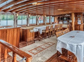 Panoramablick auf den MS Thalassa Salon mit Holzdeckenbalken, großen Fenstern, Esstischen mit weißen Tischdecken und beiger Sitzecke.