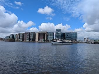 Weißes Passagierschiff Floor van Aemstel am Ufer eines breiten Gewässers mit modernen Bürogebäuden an der Amsterdamer Waterfront unter blauem Himmel.