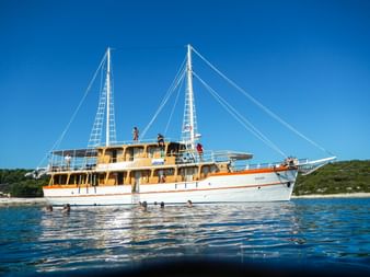 MS Dalmatino, eine weiß-gelbe Motorsegelyacht mit zwei Masten, ankert in ruhigem blauen Wasser mit schwimmenden Passagieren und grüner Küste.