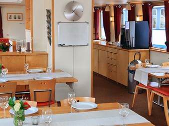 Helles Restaurant-Interieur auf der MS Fleur mit Holztischen, weißen Tischdecken, bordeauxroten Vorhängen und Fenstern mit Ausblick.