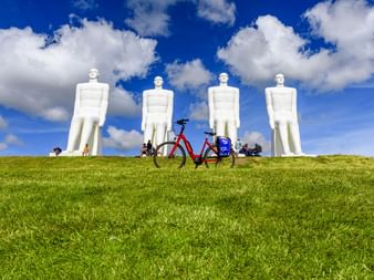 Rotes Tourenrad mit blauen Taschen auf grünem Gras vor vier großen weißen menschlichen Statuen vor blauem Himmel mit weißen Wolken.