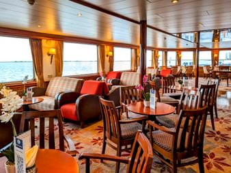 Heller Loungebereich an Bord der MS Rigoletto mit Holztischen, Stühlen, karierten Sofas und großen Fenstern mit Blick aufs Wasser.