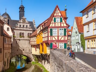 Radfahrerin auf Steinbrücke in Marktbreiter Altstadt mit bunten Fachwerkhäusern, historischem Torturm und Kanal unter blauem Himmel.