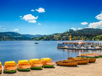 Bunte Tretboote am Sandstrand des Titisees mit Fahrgastschiff am Steg, bewaldete Hügel und blauer Himmel im Hintergrund.