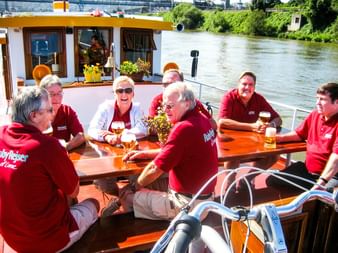 Gruppe von Radfahrern in roten Hemden sitzt an Holztischen auf dem Deck der MS Miro mit Fahrrädern und genießt Getränke an einem sonnigen Tag am Fluss.