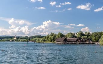 Rekonstruierte prähistorische Pfahlbauten am Bodensee mit Kirchturm in der Ferne. Blaues Wasser im Vordergrund, grüne Landschaft und Himmel.